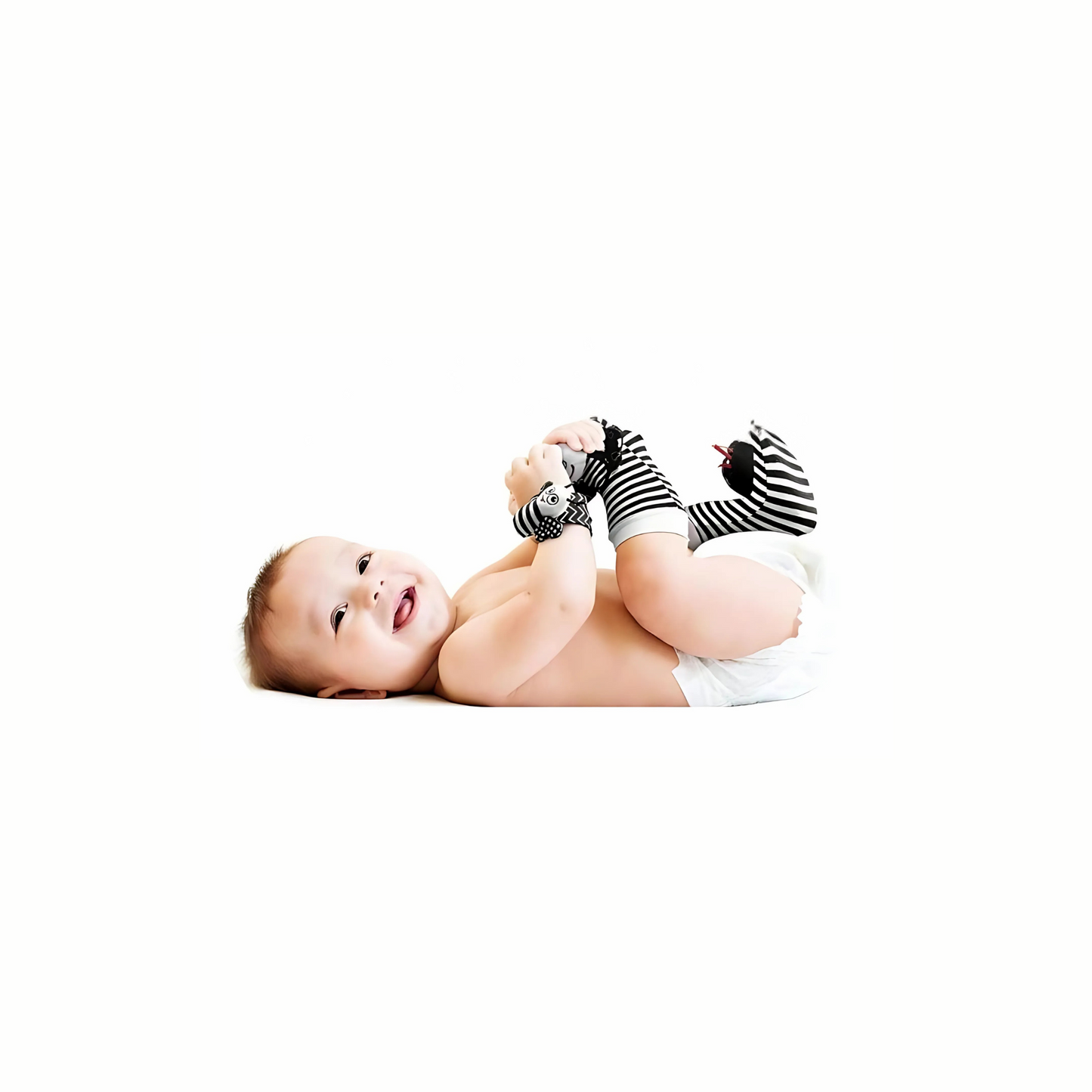 A smiling baby wearing black and white rattle socks and wristbands, lying on their back and holding their feet. This image shows the eco friendly rattle socks and wristbands being used by a child.