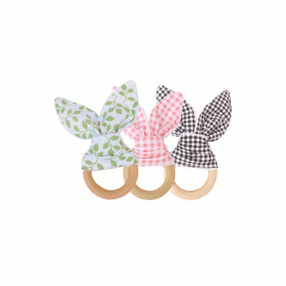 Wooden Bunny Teething Ring set showing three fabric styles: black and white gingham, pink and white gingham, and white with green leaf print. Crafted from smooth, natural wood and cotton, offering a safe and satisfying surface for teething.